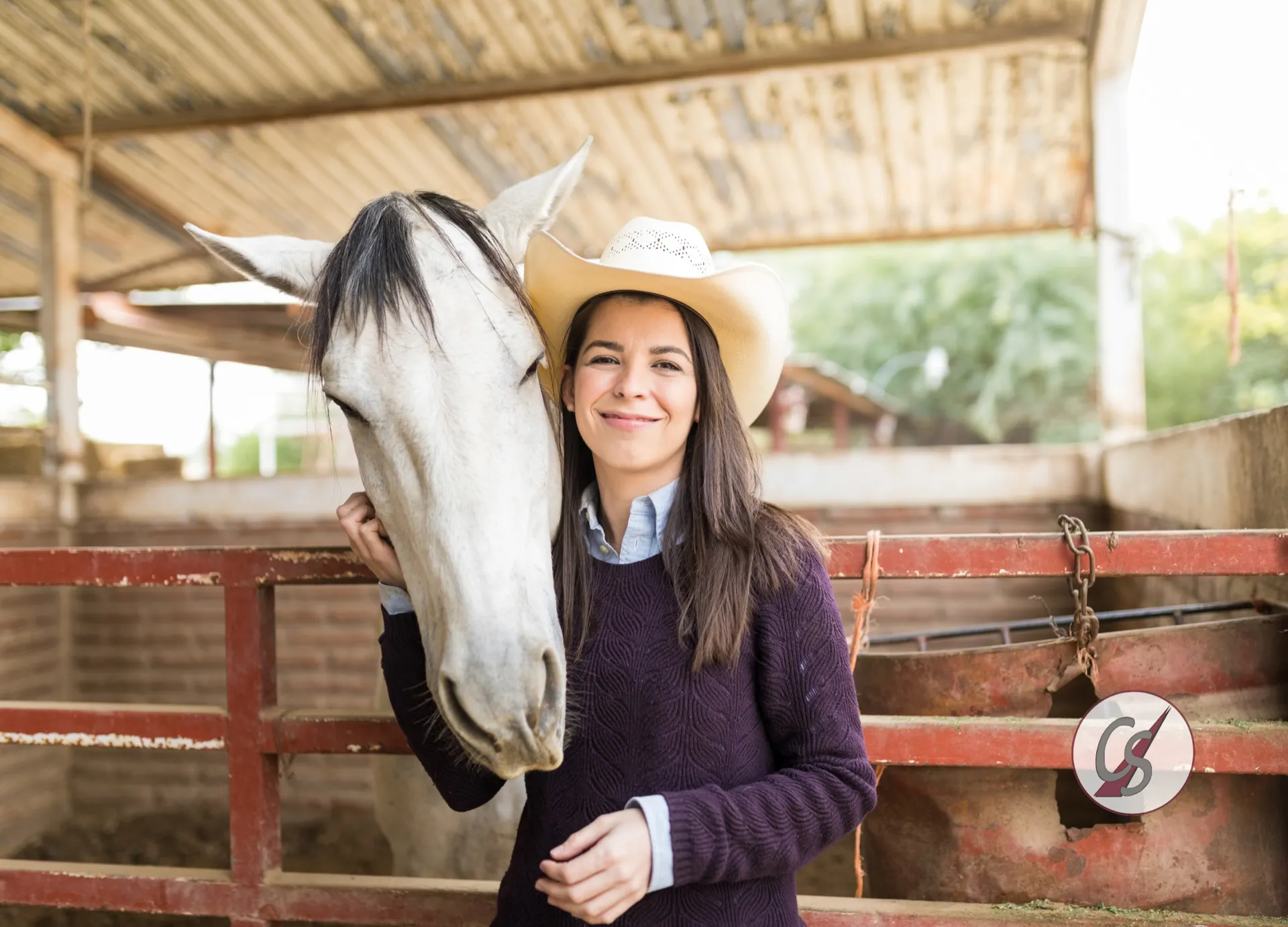 Eine lächelnde Frau mit Cowboyhut steht im Stall neben ihrem weißen Pferd
